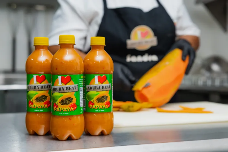Three bottles of I Love Aruba Heat papaya sauce with a chef preparing a fresh papaya in the background.