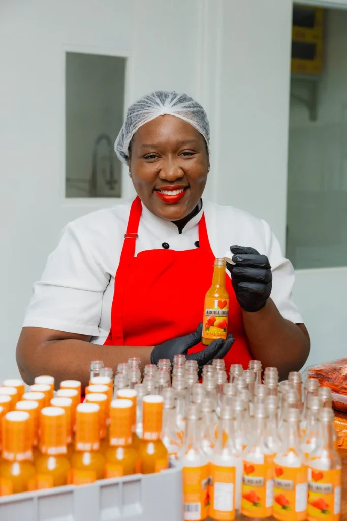 A smiling female employee in a red apron and hairnet holds up a small bottle of Aruba Heat sauce in a food production facility.